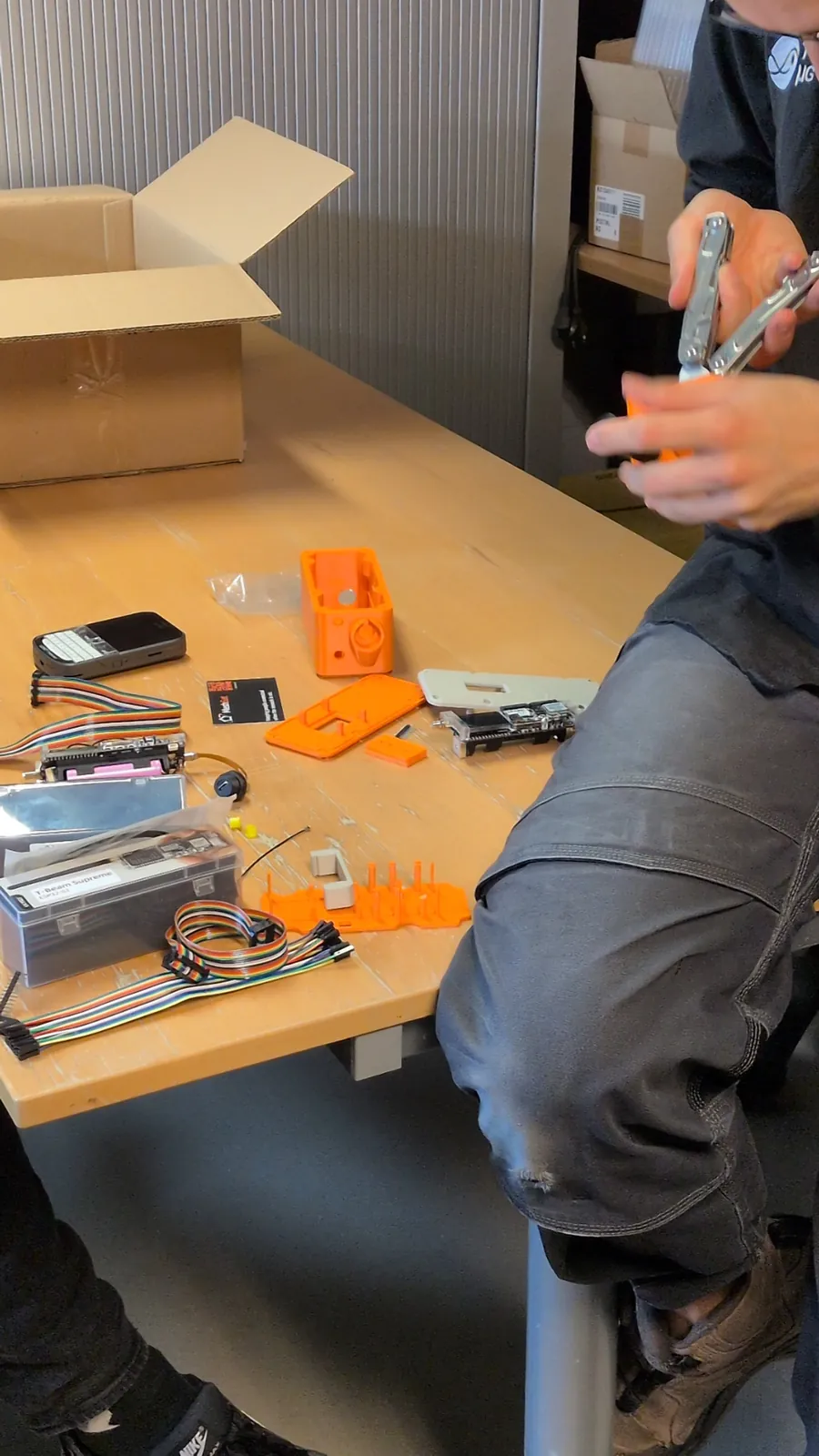 A printed orange case, a radio board and tools on a workshop table while someone works on a part.