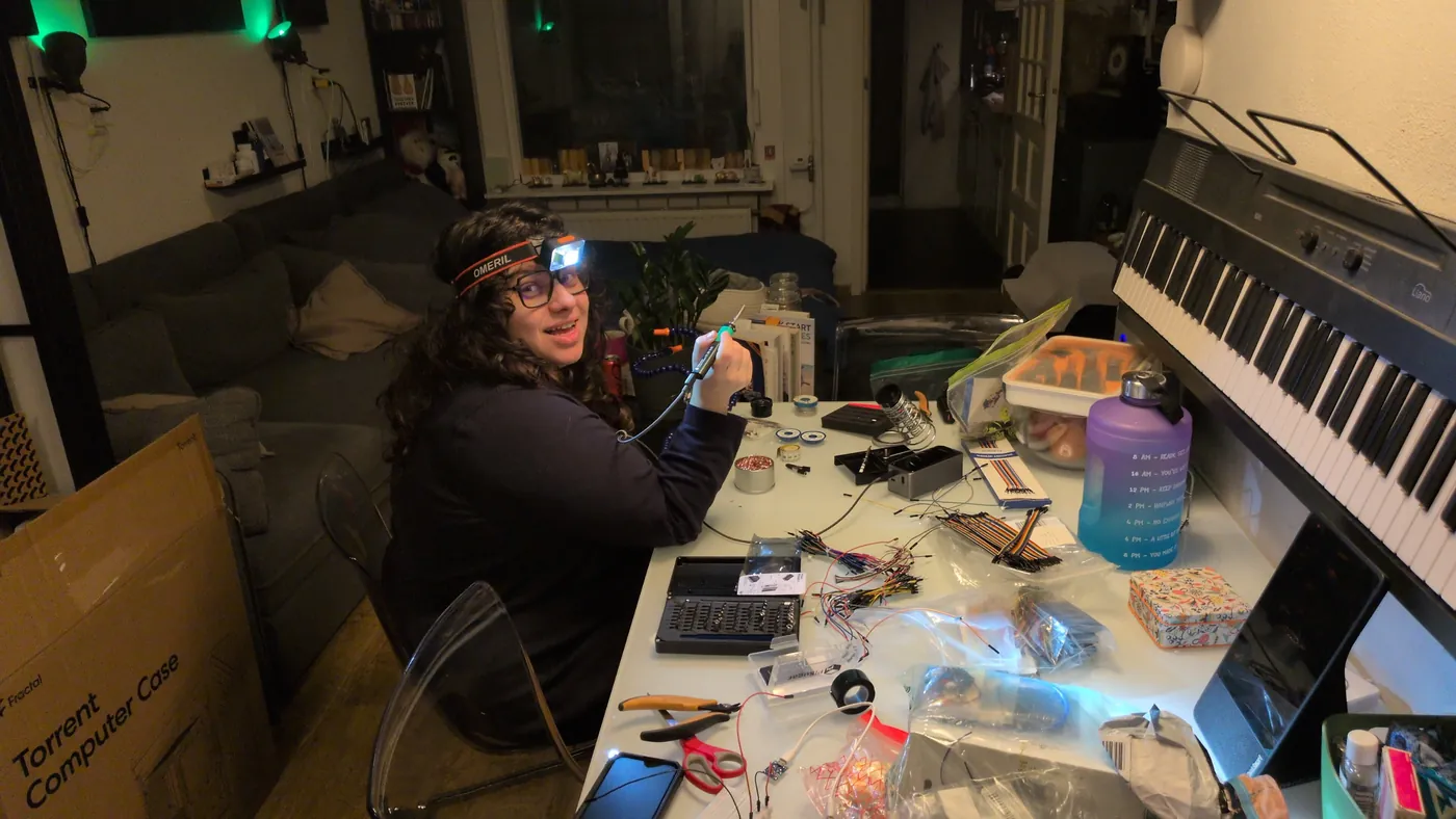 Elli at a table covered in parts, soldering under a head lamp.